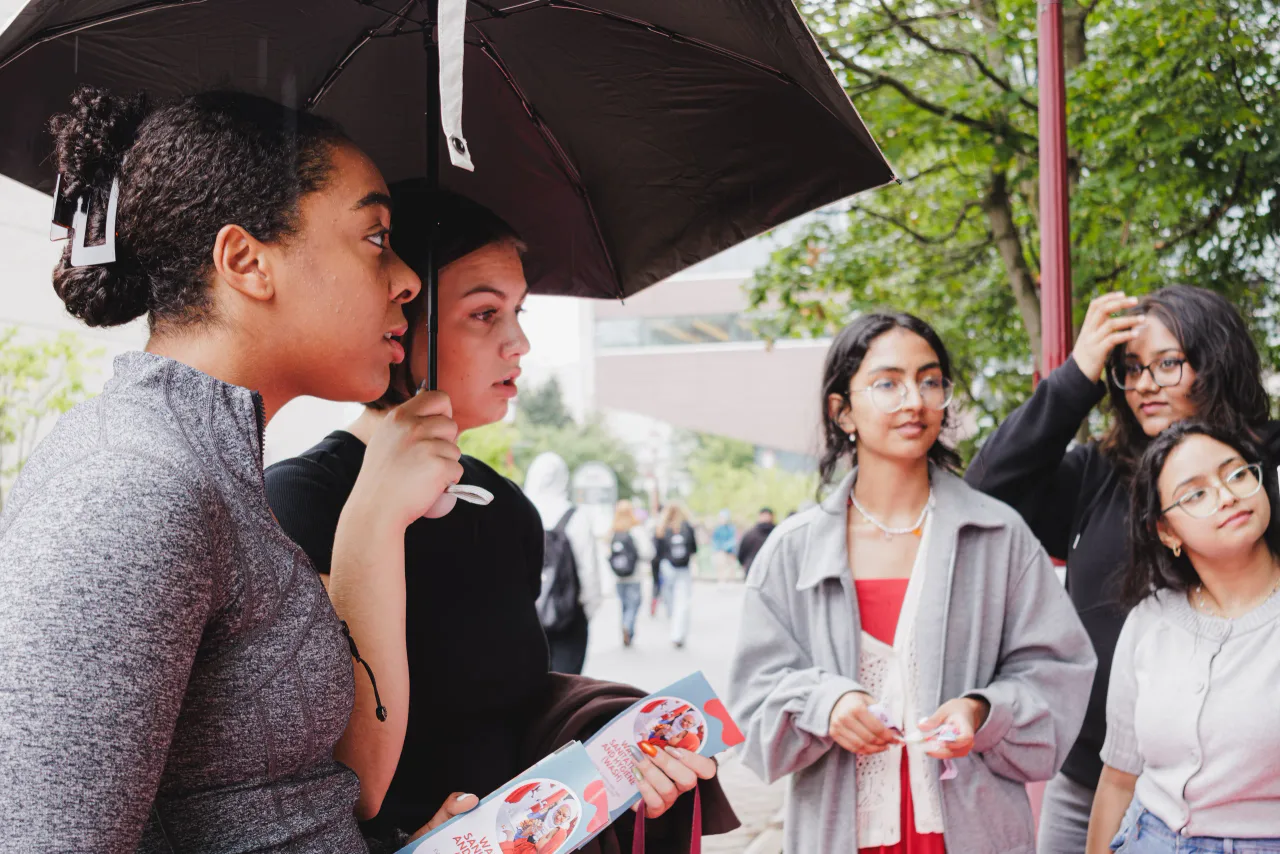 A member under an umbrella, flyer in hand, talking with four students